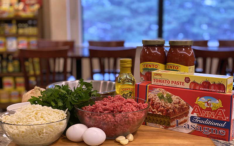 A Bowl of Shredded Mozzarella, A Bowl of Romano Cheese, A Bowl of Ricotta Cheese, A Bunch of Italian Parsley, A Bowl of Ground Beef, Two Eggs, A Yellow Onion, Three Cloves of Garlic, A Box of Anna Traditional Lasagne Noodles, A Tube of Cento Tomato Paste, and Two Jars of Cento Marinara Sauce